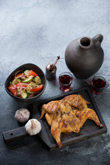 Black wooden serving tray with roasted tapaka chicken, red wine and vegetable salad. Elevated view on a grey stone background with space