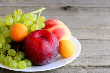 Raw fruits on a white plate and a vintage wooden background. Sweet peaches, plums, apricots, grapes branch. Natural healthy eating. Delicious summer fruits. Closeup