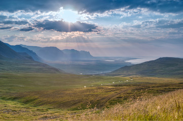 The sun's rays shine through clouds as it rises above a mountain landscape at the ocean with green fields and a meandering river in western Iceland