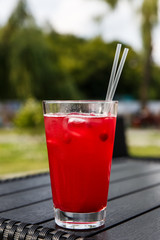 Red cranberry lemonade in a highball glass with ice cubes and 2 straws. On a dark wooden table of a city cafe terrace. Selective focus.