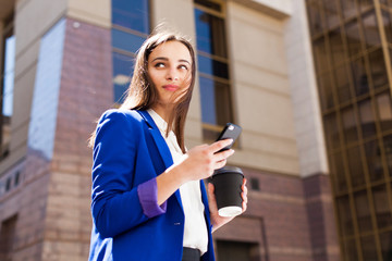 Girl in bright blue jacket stands with smartphone and coffee on the street