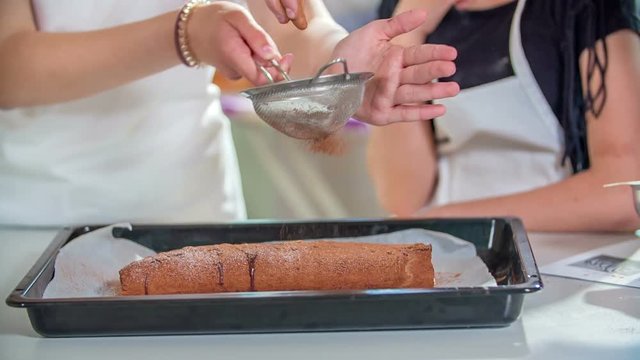 The teacher is sprinking the chocolate cake with some cocoa. One of her students is watching her.