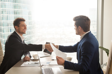 Two satisfied businessmen shaking hands at office desk after successful negotiations, business...