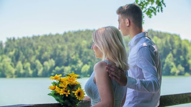 A young couple is enjoying the amazing view on the lake when a man suddenly puts his arm around his woman's back and a shoulder.