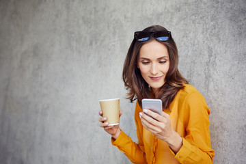 Cheerful girl browsing internet and having coffee while leaning on wall outside