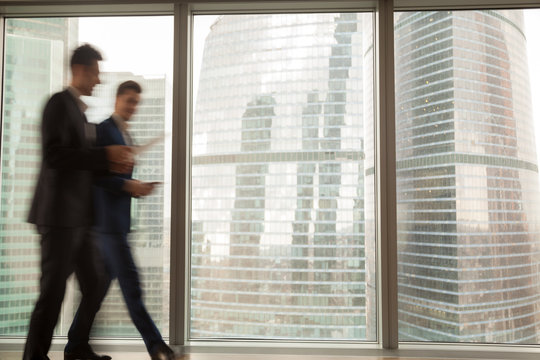 Busy businesspeople in office building, two businessmen walking along hallway, full-length window city view at background, running in hurry, business rush in lobby, working atmosphere, motion blur