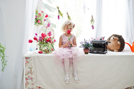 Cute little girl wearing amazing dress sitting on the windowsill next to the flowers with a tupil in her hand.