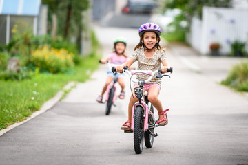 Children learning to drive a bicycle on a driveway outside.