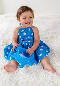 Little girl in a blue dress is sitting on the bed and laughs, playing with a toy