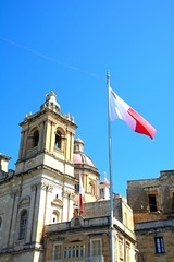 Fototapeta premium View of St Lawrence church with a Maltese flag in the foreground, Vittoriosa, Malta.