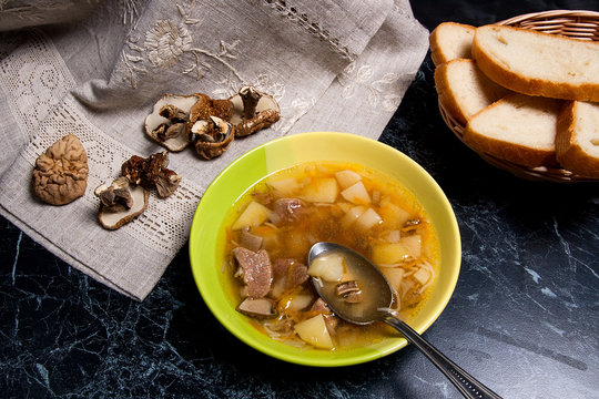Mushroom soup in green plate with metal spoon, dried wild mushrooms and basket of bread on a black stone background.