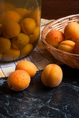 Fresh organic harvested apricots in basket and big glass jar with canned apricot compote on dark marble..