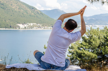 Young beautiful smiling man doing yoga exercises on kerimat on top of mountain looking at the sea resting and meditating