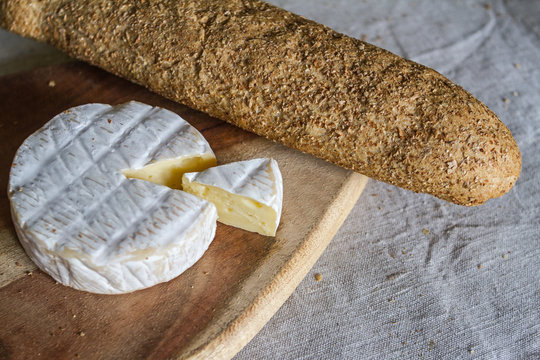 The head of the Swiss Camembert cheese and a triangular piece of cheese on a wooden textured board and a grain baguette on a woven canvas background.