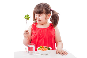 Girl prefers healthy food, sits at the table near the plate with vegetables. Child with broccoli isolated on white background