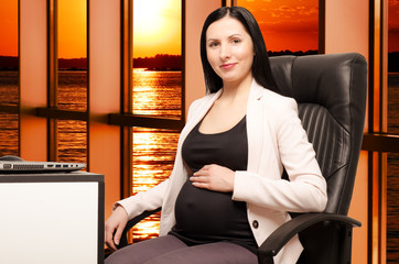 Beautiful young pregnant woman sitting on workplace in the office at sunset