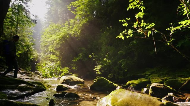 Silhouette of a man with a backpack, crosses a mountain river or stream. Beautiful forest with rays of the morning sun. Adventure and extreme tourism