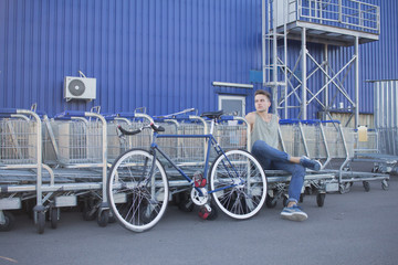 Young handsome male stitting in shopping basket cart, hipster bicycle rider posing on mall parking  