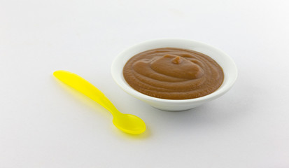 A bowl of prune and apple mixture baby food with a yellow plastic spoon to the side atop a white linen tablecloth.