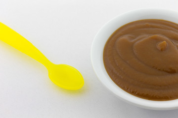 Close view of a bowl of prune and apple mixture baby food on a white linen tablecloth with a yellow spoon to the side.