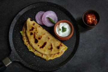 Aloo Paratha - Indian potato stuffed wheat bread served with Raita and mango pickle on dark moody background, Top down view