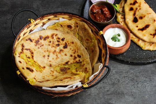 Alu paratha - North Indian Potato stuffed bread served in a basket with raita and mango pickle on dark moody grey background
