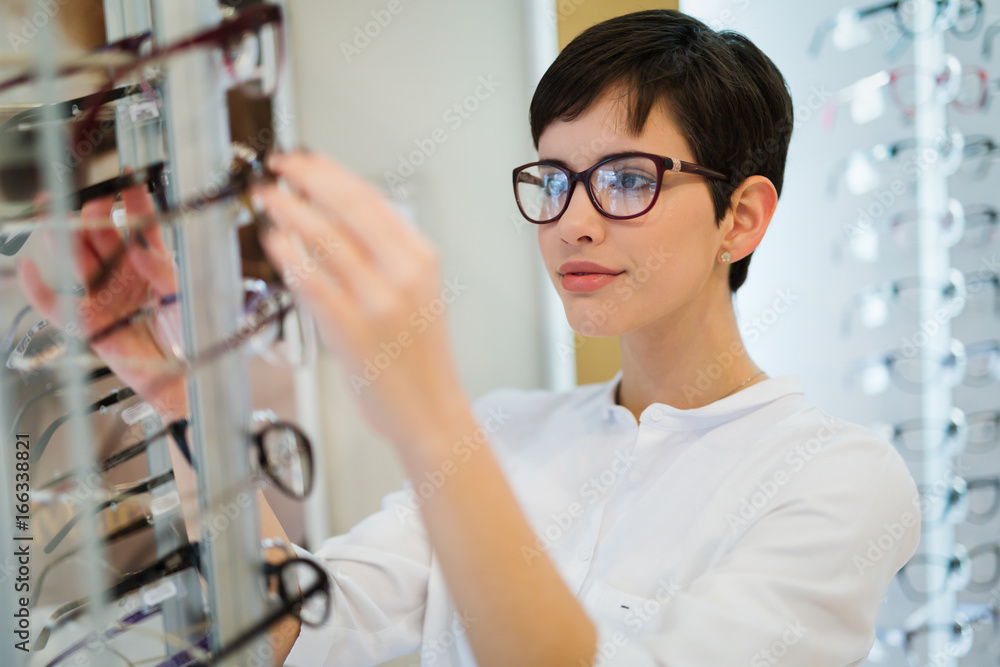 Poster pretty young woman is choosing new glasses at optics store