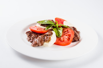 Meat salad. Fried beef with mozzarella, tomato and basil on a white plate on a light background. Close
