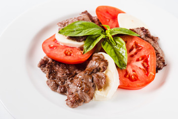 Meat salad. Fried beef with mozzarella, tomato and basil on a white plate on a light background. Close