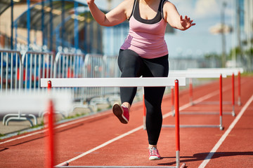 Plump young woman running down stadium racetrack while taking part in hurdle race