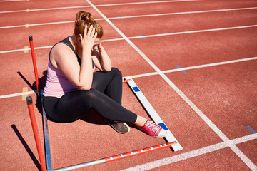 Desperate plump young woman seated on racetrack inside fallen obstacle