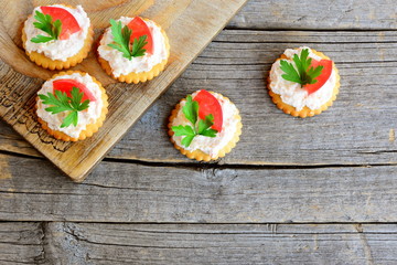 Canape cooked from salty crackers with spicy soft cheese, tomato slice and green parsley leaf. Spicy cheese canape on an old wooden background. Quick aperitif recipe. Top view