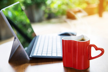 Red cup of coffee with laptop in the background place on wooden table in garden , warm light effect