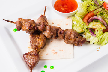 Shish kebab on bamboo skewers lies on a pita. Near sauce and fresh vegetable salad. White background (close)