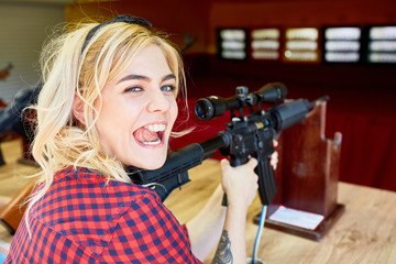 Portrait of cheeky blond girl holding big rifle at shooting stand in amusement park