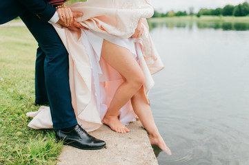 Fototapeta na wymiar Unrecognizable bride in expensive stylish wedding dress pushing her slim naked beautiful leg in river. Unknown groom in suit holding his wife hand and body. Wedding day celebration. Bridal walking.