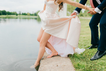 Fototapeta na wymiar Unrecognizable bride in expensive stylish wedding dress pushing her slim naked beautiful leg in river. Unknown groom in suit holding his wife hand and body. Wedding day celebration. Bridal walking.