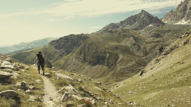 Woman trekking in idyllic mountain landscape on footpath crossing blooming green meadow set amid high altitude rocky mountain range and peaks. Summer adventures on the Italian Alps. Slow motion.
