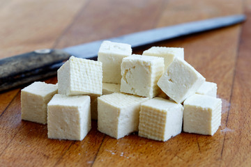 Cubes of cut white tofu and rustic knife on wooden chopping board.