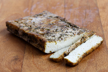 Block of marinated tofu with herbs and two tofu slices on wooden chopping board.