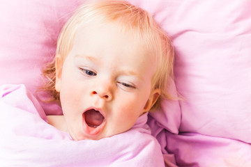 An awakened baby in a bed on a pink cushion yawns