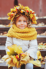 cute adorable toddler girl vertical portrait with bouquet of autumn leaves and wreath walking outdoor in park or forest, wearing warm knitted yellow snood