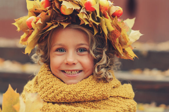 cute adorable toddler girl vertical portrait with bouquet of autumn leaves and wreath walking outdoor in park or forest, wearing warm knitted yellow snood