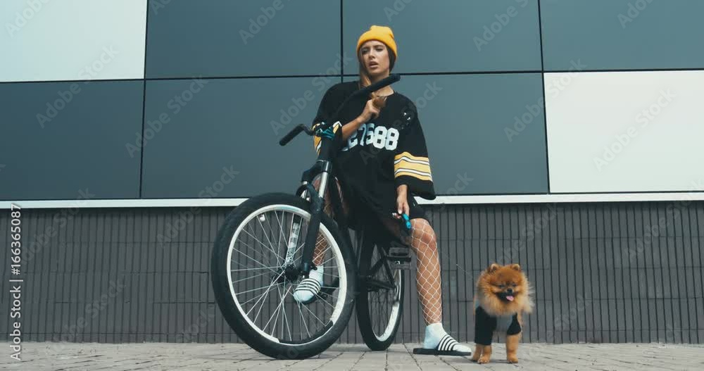 Wall mural Young stylish Caucasian female posing on a bicycle with her Pomeranian Spitz dog puppy outdoors. 4K UHD RAW edited footage