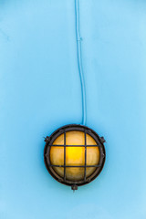 Ship yellow deck lamp surrounded by a metal rusted frame fixed to a painted light blue wooden wall and similarly painted electric cable