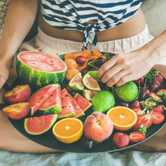 Summer healthy raw vegan clean eating breakfast in bed concept. Young girl wearing striped home shirt sitting and taking fruit from tray full of fresh seasonal fruit, square crop