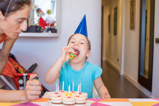 three years old blonde child, with blue cone hat, ready to blow a party blower, next to woman and birthday cake with candles on colorful tablecloth at home
