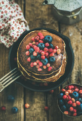 Sweet breakfast with pancakes with fresh forest berries and honey in cast iron pan over rustic wooden background, top view