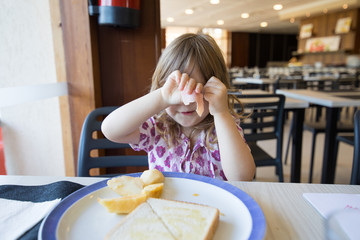 portrait of blonde caucasian child three years old, eating, hiding face with hands with slice of ham, and bread toast in dish, sitting indoor in restaurant
