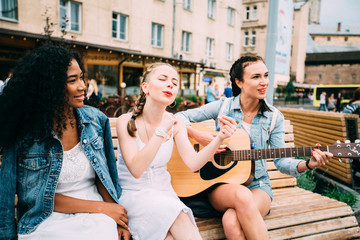 Cheerful multiethnic hipster teenagers girlfriends playing guitar and singing.